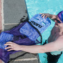 Swimmer at the edge of a pool placing a blue marbled Maru Swim kickboard into a purple mesh swim bag. The kickboard features cutout handles and the Maru logo, ideal for swim training exercises.