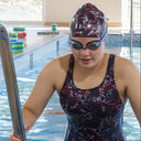 Person in a swimsuit and swim cap standing by a pool with safety signs on the wall.